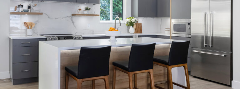 Spaces12-home-renovation-port coquitlam-1 Modern kitchen with gray cabinets, white countertops, a stainless steel refrigerator, and three black stools at a central island.