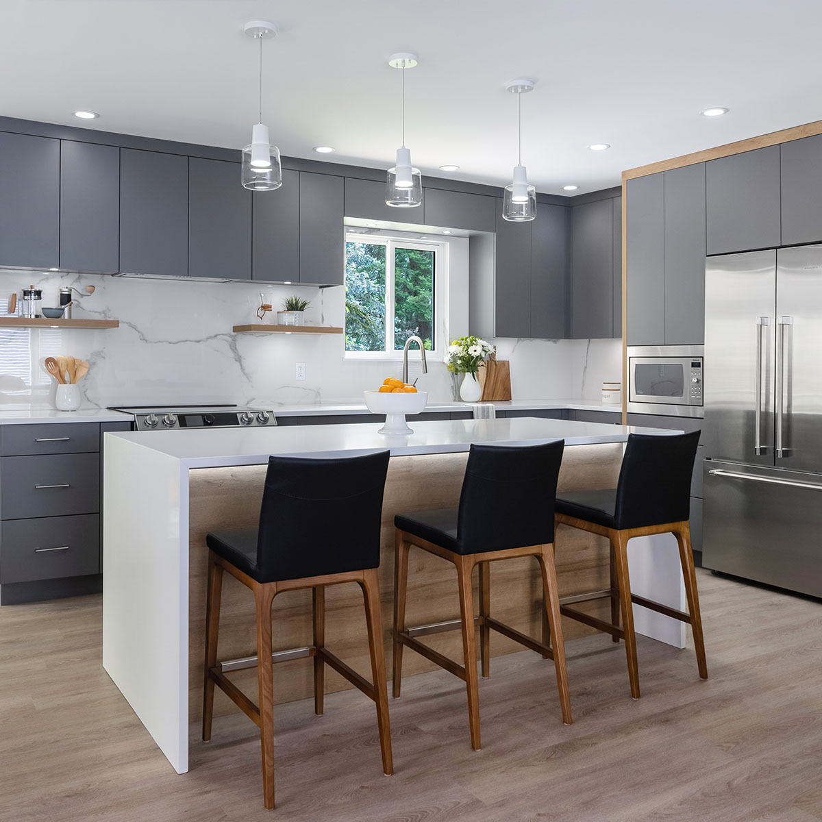 Modern kitchen with gray cabinets, white countertops, and stainless steel appliances. An island with a wooden base and three black bar stools is centered under three pendant lights.